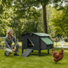 Sfeerfoto Nestera kunststof kippenhok groot op poten schuin vooraanzicht, in een achtertuin op het gras. Naast het kippenhok zit een vrouw gehurkt en voor het kippenhok loopt een kip.